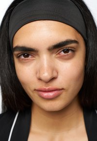 Svart elastisk hårband som bärs på mörkt hår, närbild av ett ansikte som visar hudens textur, bruna ögon och neutral läppfärg.