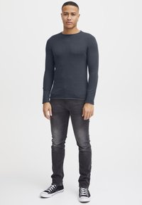 Camicia a maniche lunghe, aderente, grigia scura con colletto rotondo, abbinata a jeans grigi scuri e sneakers nere con suola bianca.