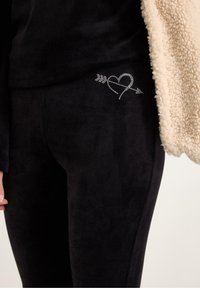 Schwarze Fleece-Leggings mit einem Herz- und Pfeil-Design aus klaren Strasssteinen auf dem linken Oberschenkel, kombiniert mit einer cremefarbenen, flauschigen Strickjacke.