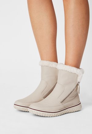 Bottes d'hiver beiges mi-mollet avec doublure en fourrure, fermeture éclair latérale et semelle texturée portées sur des jambes nues sur fond blanc.