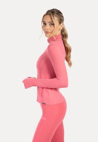 Langærmet lyserød sportstop med høj krave, tætsiddende design og tommelhuller. Glat stof og diskret logodetalje ved kanten.