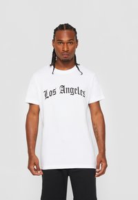 Bílý bavlněný tričko s nápisem "Los Angeles" tučným černým písmem na hrudi. Krátké rukávy a volný střih s hladkou texturou.
