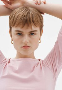 Top de manga corta, en un suave color rosa, con un escote relajado que presenta un ligero gather en los hombros. La modelo tiene el cabello corto y lleva pendientes de aro dorados.