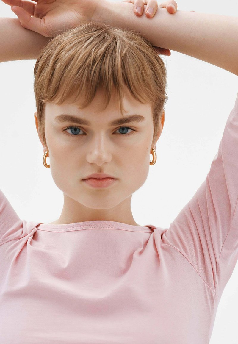 Top de manga corta, en un suave color rosa, con un escote relajado que presenta un ligero gather en los hombros. La modelo tiene el cabello corto y lleva pendientes de aro dorados.