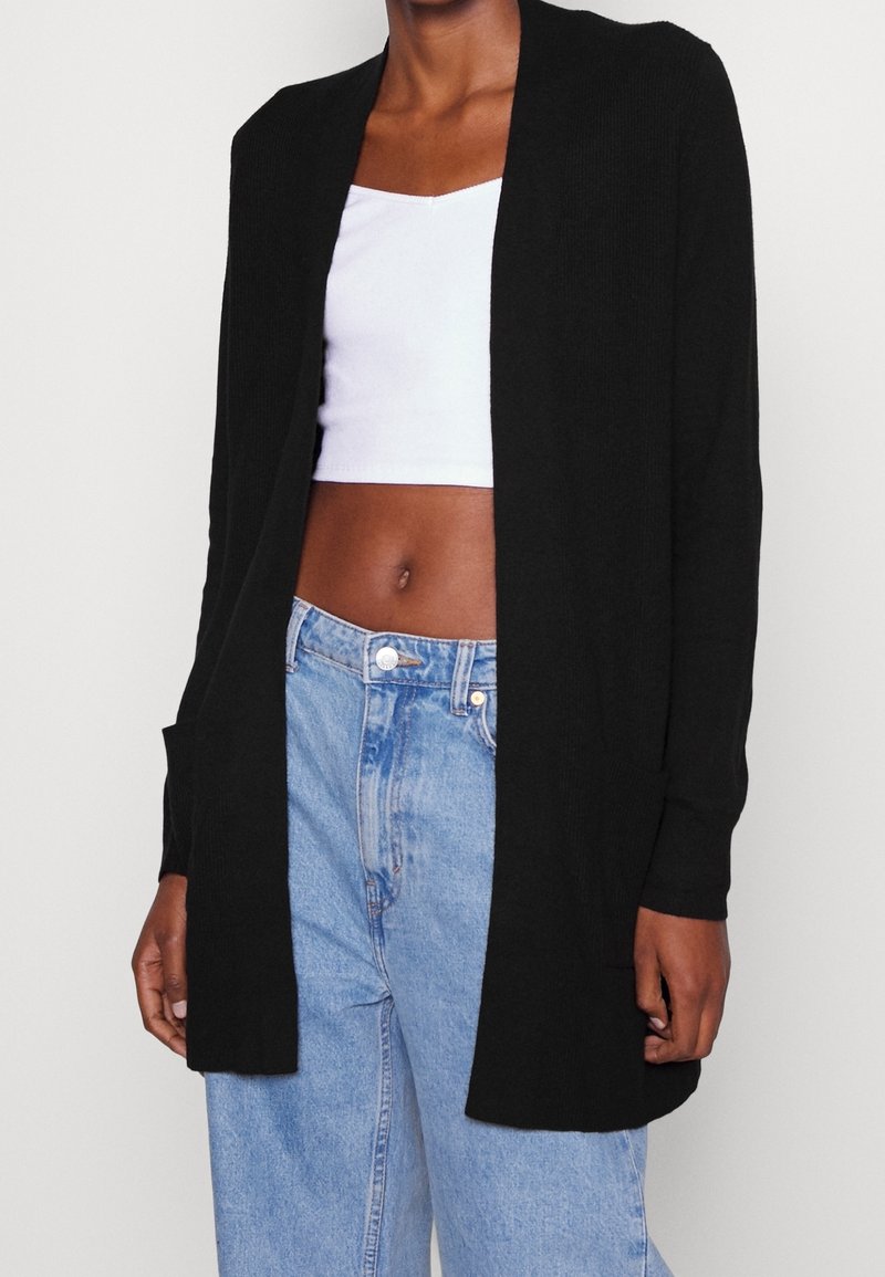 Cardigan noir ouvert devant avec manches longues, texture côtelée et deux poches latérales, associé à un jean bleu taille haute et un haut blanc.