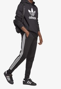 Svart hoodie med vitt Adidas-logotyp, kombinerad med svarta sweatpants med tre vita ränder på sidorna och svarta sneakers.