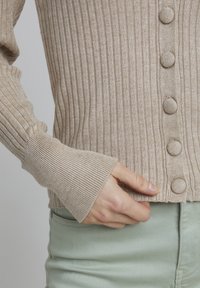 Beige ribbet cardigan med runde stofknapper, med en blød tekstur og tætsiddende ærmer, båret over lysegrønne bukser.