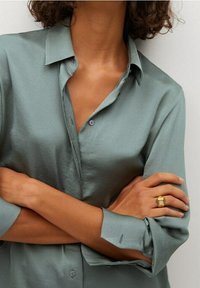 Personne aux bras croisés portant une chemise à boutons vert clair et une bague dorée à texture sur l'annulaire de la main droite.