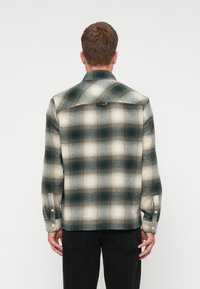 Camisa de franela a cuadros en tonos verde, crema y gris. Presenta mangas largas, cuello puntiagudo y botones de presión. Tejido suave y texturizado.