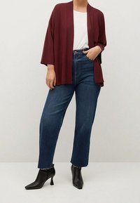 Bordeaux gestrickte Strickjacke über einem weißen Hemd, kombiniert mit hochgeschnittenen blauen Jeans und schwarzen Stiefeletten mit Absatz. Einfaches Design und Passform.