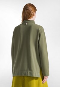 Olijfgroene sweatshirt met een kraag, drop schouders en zijsplitten, gecombineerd met een textuur gele rok. Zachte stof met minimale details.