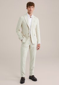 Világos bézs színű vászon öltöny, testhezálló szabással, egysoros zakóval, passzoló nadrággal, és sötétbarna mokaszinokkal. Fehér ing az alatt.