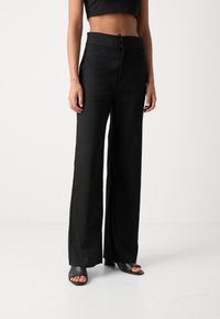 Pantalones negros de pierna ancha con una tela lisa y brillante, cintura alta y un diseño elegante; combinados con mules negros de tacón y punta abierta.