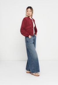 Woman wearing a white top, red corduroy jacket, wide-leg blue jeans, and beige sneakers, standing with hands in pockets against white background.