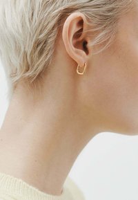 Pendiente de aro dorado con un diseño redondeado y abierto. Textura suave con un acabado pulido, llevado en una oreja perforada. Cabello corto y ligero visible.