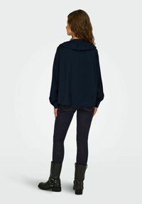 Chemise oversize bleu marine avec capuche, manches longues et ourlet arrondi. Associée à un jean foncé et des bottines noires à boucles.