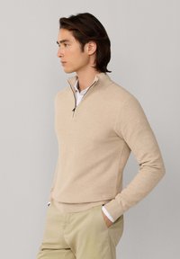 Muž nosiaci béžový sveter so zipsom do štvrťky cez bielu košeľu a béžové nohavice, stojaci s rukami v vreckách na jednoduchom pozadí.