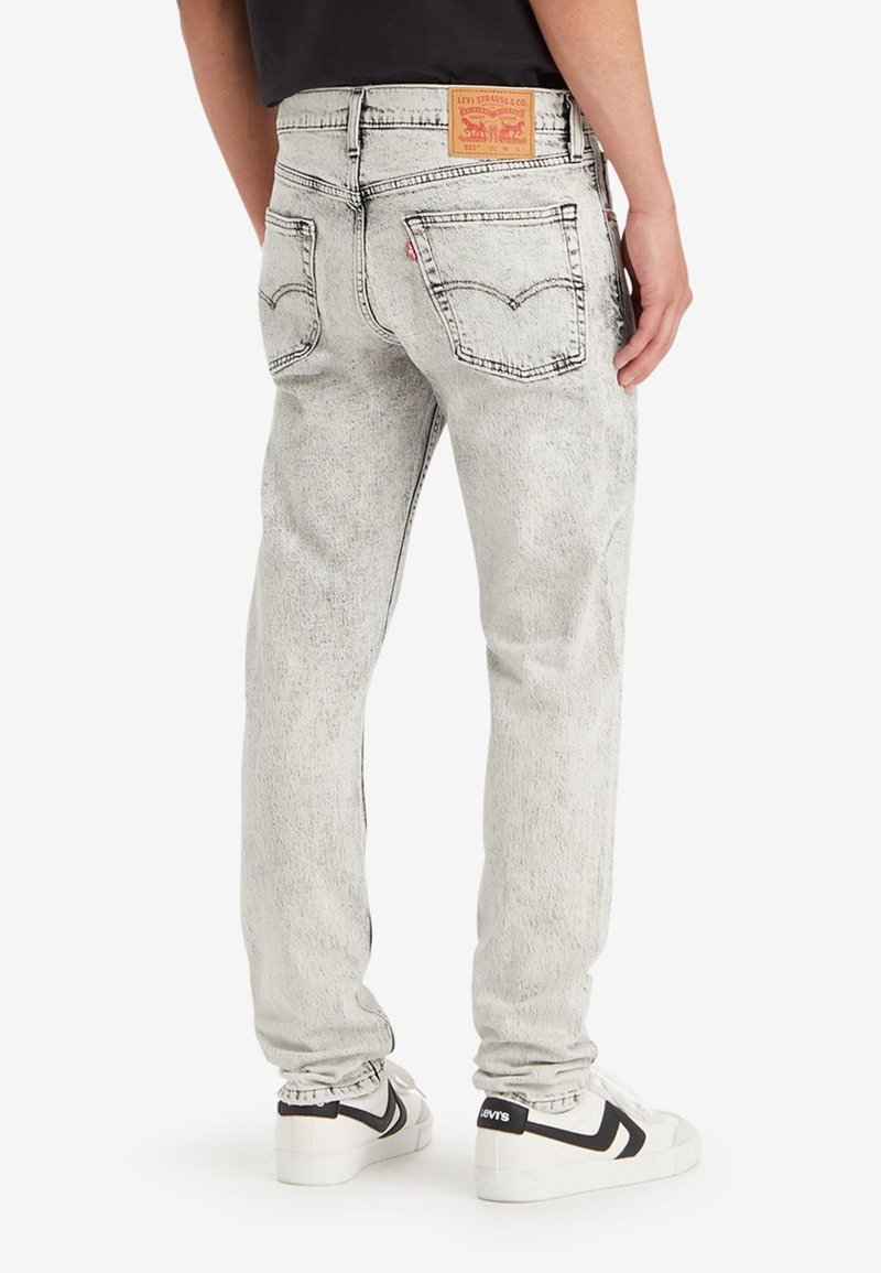 Personne portant un jean Levi’s gris clair acidulé et des baskets blanches avec des détails noirs, debout dos à la caméra.