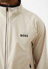 Hellbeige Zip-Jacke mit hohem Kragen, ausgestattet mit schwarzem Metall und einem schwarzen "BOSS"-Logo auf der linken Brustseite. Glatte Textur.