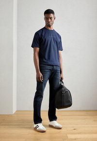 Tricou din bumbac navy cu mâneci scurte, blugi din denim, adidași albi cu accente negre și o geantă de voiaj din piele neagră.