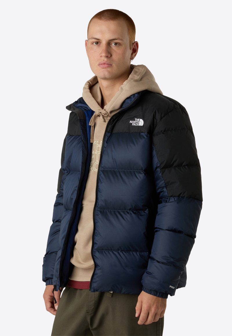 Dunkelblaue Steppjacke mit schwarzen Schultern, ausgestattet mit einem Reißverschluss vorne, elastischen Bündchen und einem Logo auf der linken Brust. Weicher, gesteppten Textur.