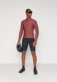 Fietsoutfit bestaande uit een strakke bordeauxrode lange mouw top met een rits aan de voorkant en zwarte shorts. Accessoires zijn een zwarte helm en een zonnebril.
