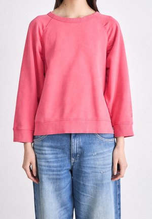 Vrouw draagt een felroze sweatshirt met lange mouwen en lichtblauwe verweerde spijkerbroek, staand tegen een effen witte achtergrond.