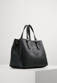 Borsa tote in pelle nera con texture a granuli, due manici e dettagli in metallo dorato. Il nome del marchio è impresso in oro sul davanti.