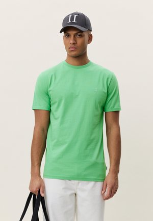 Mann iført grønn kortermet skjorte og grå caps med "II"-logo, holder svarte veskehåndtak og står foran en ensfarget lys bakgrunn.