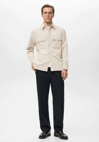 Camisa de botón beige claro con dos bolsillos en el pecho, llevada sobre una camiseta blanca. Combinada con pantalones oscuros y zapatos negros, sobre un fondo blanco.