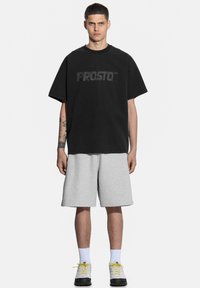 Czarny bawełniany t-shirt z logo "PROSTO", w zestawieniu z szarymi szortami. Model nosi białe skarpetki i szare obuwie z żółtymi sznurówkami. Prosty design.