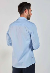 Chemise légère bleue à manches longues avec un col pointu, texture douce, ourlet arrondi et un petit logo près du bas.