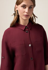 Burgundy skjorta med knappar, två bröstfickor, svarta knappar och en slät textur. Noterbar kristallaccessoar i mitten.