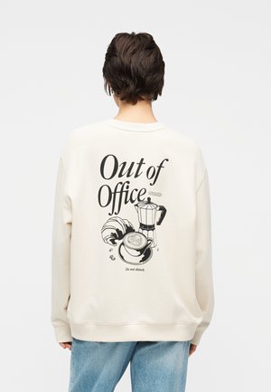 Személy krémszínű pulóvert visel, amelynek hátulján "Out of Office" felirat és fekete kávéfőző, croissant és kávéscsésze illusztráció látható.