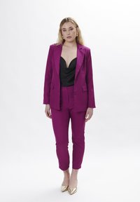 Traje magenta con chaqueta ajustada y pantalones entallados, combinado con una blusa negra de cuello cisne y zapatos de tacón alto dorados. Textura de tela suave.