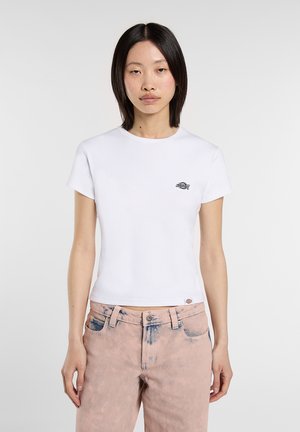 Mujer joven con cabello lacio y negro, vistiendo una camiseta corta de manga corta blanca y jeans de lavado ácido rosa claro, de pie frente a un fondo blanco.