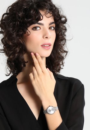 Joven mujer con cabello oscuro y rizado, vistiendo una camiseta negra y un reloj de pulsera plateado, tocándose la barbilla con los dedos.