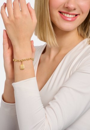 Donna sorridente con capelli corti biondi che indossa un top bianco, mostrando un bracciale a catena d'oro con un ciondolo rettangolare al polso.