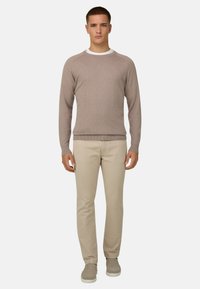 Homme vu de face portant un pull beige par-dessus une chemise blanche, un pantalon beige clair et des chaussures légères beiges sur un fond uni.