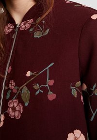 Veste bomber bordeaux avec broderies florales en rose et vert, dotée d'une fermeture éclair argentée et d'un col côtelé. Texture lisse.