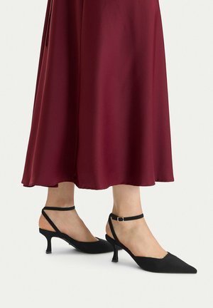Mujer con zapatos negros de tacón bajo puntiagudos con tiras en el tobillo y falda midi burdeos sobre un fondo liso.