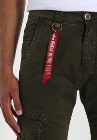 Pantalones cargo verde oliva con un acabado texturizado, que cuentan con un cierre de botón metálico y una etiqueta de tela roja que dice "RETIRAR ANTES DEL VUELO."