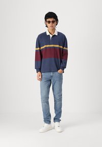 Langermet polo i marineblå med horisontale striper i burgunder og gul. Kombinert med lyseblå jeans og hvite sneakers.