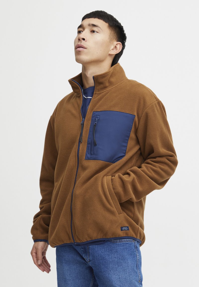 Brun fleecejakke med en blå nylon brystlomme og svart glidelås. Har høy krage og blå aksenter, kombinert med bluesjeans.