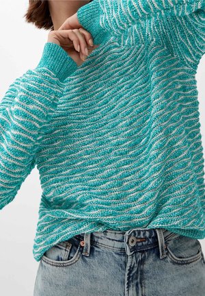 Pull en maille texturée turquoise et blanc avec des poignets côtelés, porté rentré dans un jean taille haute délavé clair, main ajustant le col.