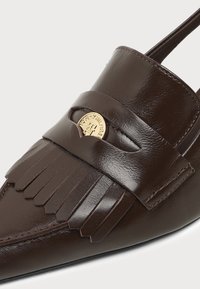 Σκουρόκαφέ δερμάτινο loafers με κρόσια και στρογγυλό χρυσό έμβλημα στο λουράκι που δείχνει το λογότυπο του εμπορικού σήματος.