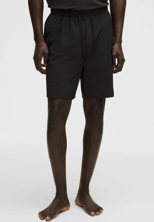 Man draagt zwarte knielange shorts met een elastische tailleband en trekkoord, staand op blote voeten tegen een effen achtergrond.