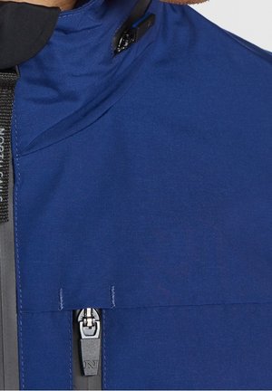 Gros plan sur une veste bleue avec fermeture éclair noire et sangle noire arborant un logo "N" visible, portée par une personne.