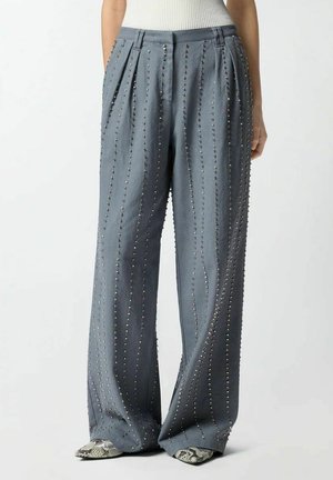 Donna che indossa pantaloni grigi a gamba larga con decorazioni verticali di borchie e scarpe a punta con motivo in pelle di serpente.