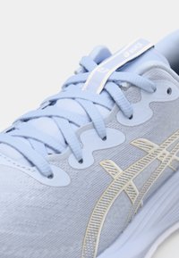 Baskets de sport bleu clair avec une tige texturée, des lacets fins et des accents dorés. Inclut un logo et des éléments de design élégants.
